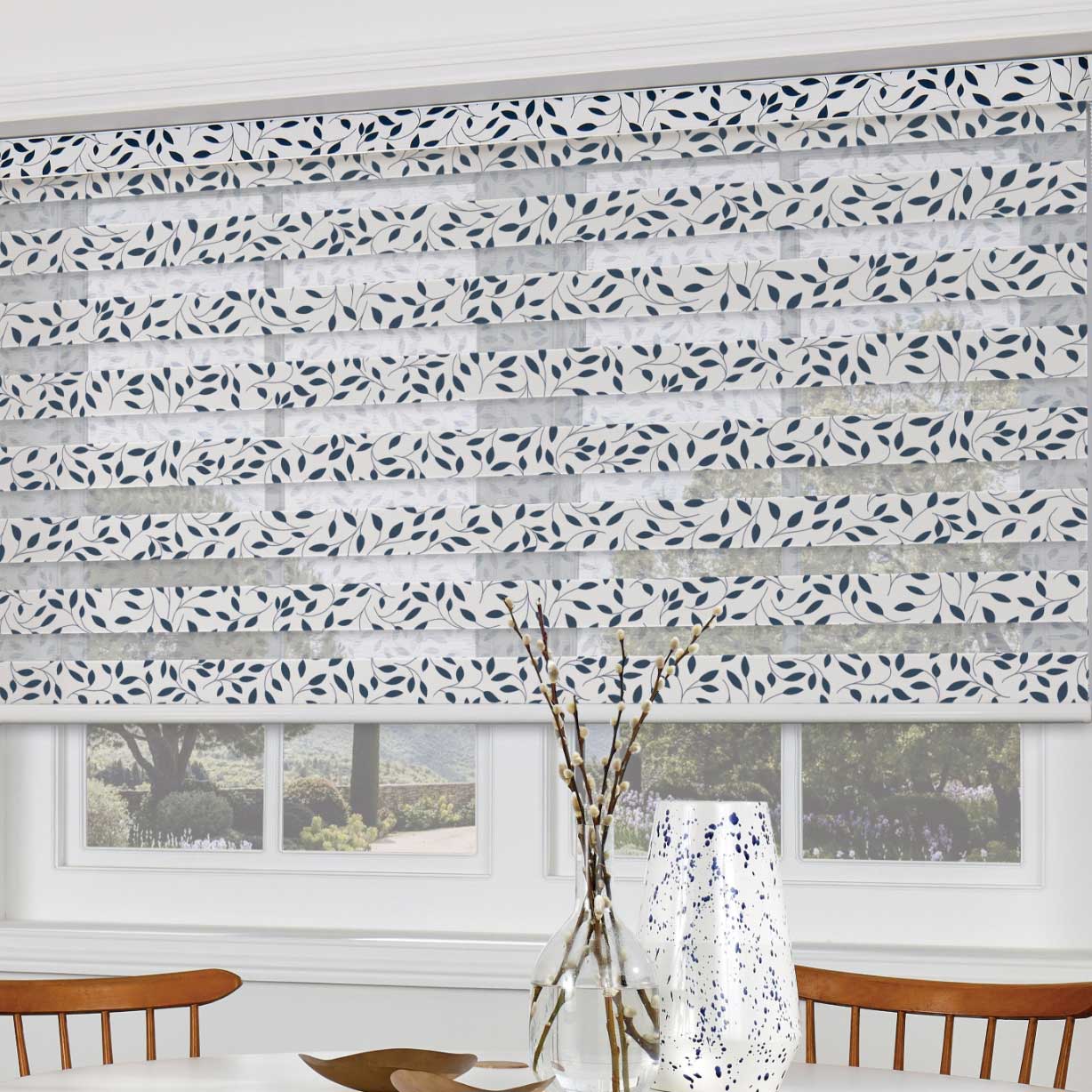 Leaf patterned white Day Night blind on bright kitchen window next to a dining table and wooden chairs